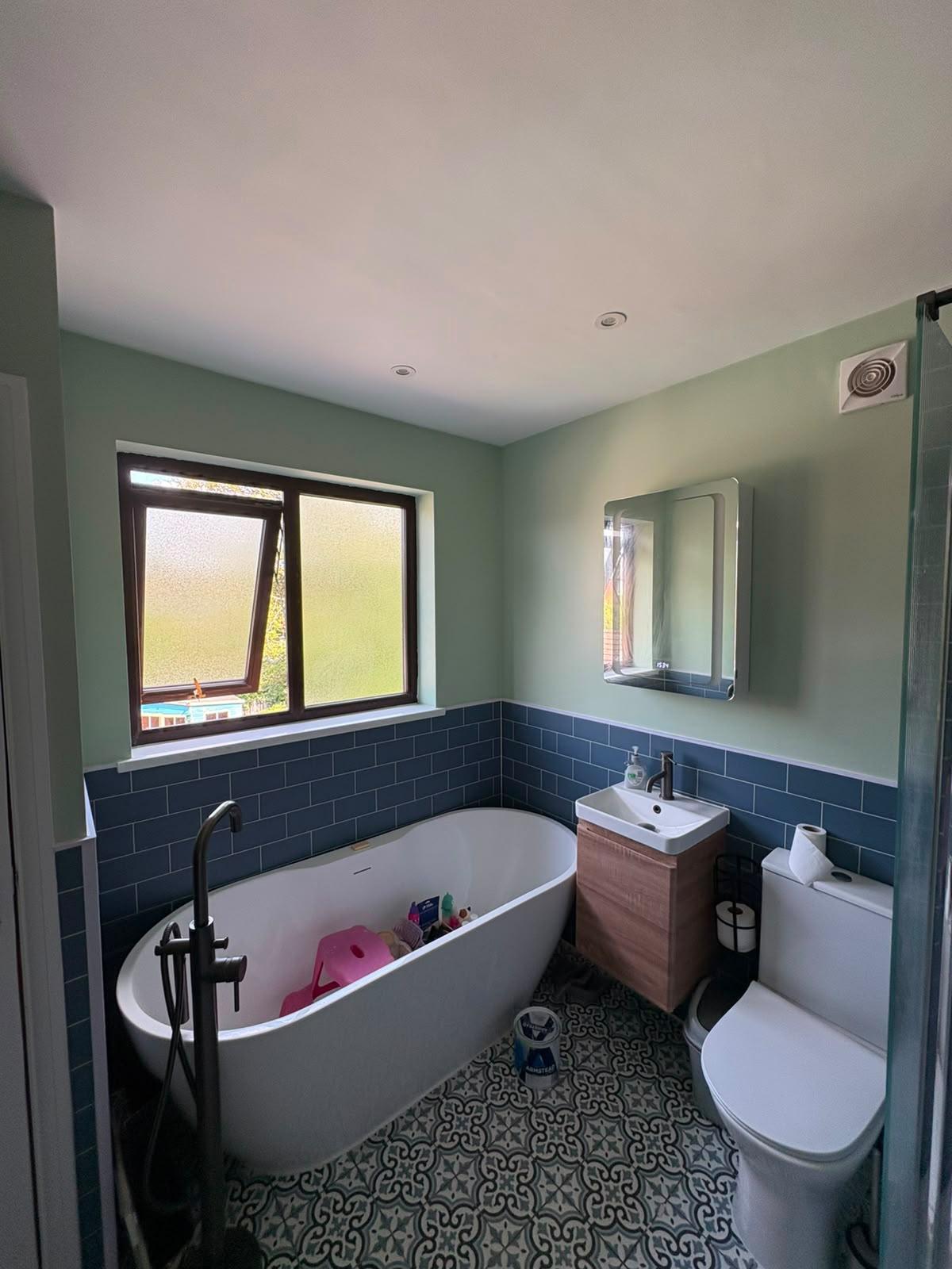 Bathroom painted eucalyptus green above dark blue metro tiles, with a freestanding bath and black taps